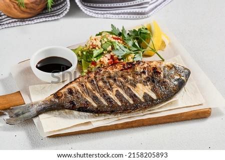 Grilled dorado with garnish on concrete background. Aesthetic composition with bbq fish and vegetables. Healthy food - roasted dorado. Fish dish in minimal style. Whole grilled fish