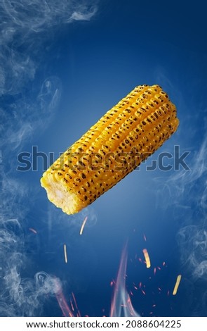 Grilled ear of corn hovering over flames in sparks and haze on blue background