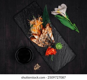 Grilled eel being served on a stone plate with ginger, wasabi, vegetables and a bowl of soy sauce, on a dark wooden table