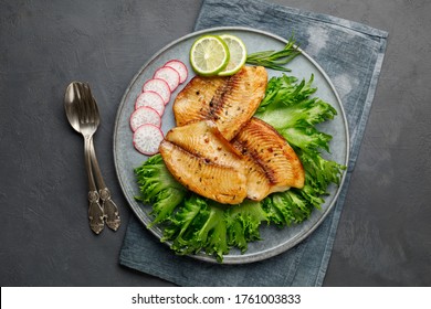 Grilled Fish Sea Bass Fillet With Green Lettuce In A Plate On A Black Stone Background. Above View.