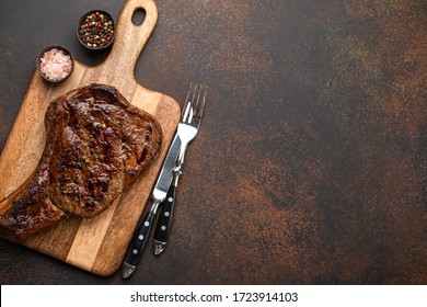 Grilled or fried prime marbled meat steak Ribeye served on wooden cutting board with cutlery. Top view of juicy cooked beef steak on rustic brown concrete background with space for your text - Shutterstock ID 1723914103