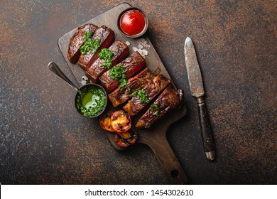 Grilled or fried and sliced marbled meat steak with fork, tomatoes as a side dish and different sauces on wooden cutting board, top view, close-up, stone rustic background. Beef meat steak concept 