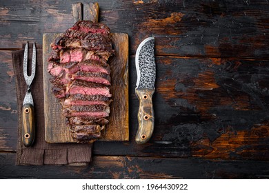 Grilled or fried and sliced marbled meat steak rib eye set, on wooden serving board, with meat knife and fork, on old dark wooden table background, top view flat lay, with copy space for text