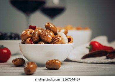 grilled green olives in oil, pitted, with red hot pepper and garlic in a white bowl, still life with tomatoes, glasses of red wine, wooden skewers for olives, side view - Powered by Shutterstock