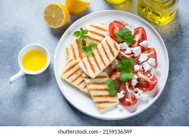 Grilled halloumi cheese with red tomatoes and feta on a white plate, high angle view on a light-blue stone background, horizontal shot