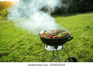 Grilled meat in grill. Roasting steaks and vegetables bbq in a round grill in a meadow. Summer picnic outdoors. - Powered by Shutterstock