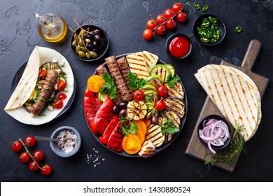 Grilled meat kebabs, vegetables on a black plate with tortillas, flat bread. Slate stone background. Top view.