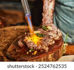 Grilled meat sizzling as a chef uses a blowtorch to add flavor, creating a smoky, caramelized texture on a wooden board.