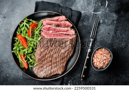 Grilled medium rare flank beef steak with salad in a plate. Black background. Top view.
