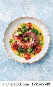 Grilled Octopus Tentacle With Broccoli, Cherry Tomatoes And Baked Baby Potatoes In White Porcelain Plate Over Blue Concrete Background. Overhead View, Copy Space 