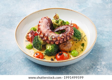 Grilled octopus tentacle with broccoli, cherry tomatoes and baked baby potatoes in white porcelain plate over blue concrete background. Side view, close up