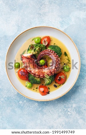 Grilled octopus tentacle with broccoli, cherry tomatoes and baked baby potatoes in white porcelain plate over blue concrete background. Overhead view, copy space 