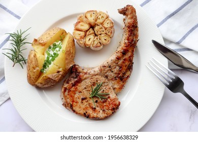 Grilled Pork Chop Steak With Baked Potato And Roasted Garlic In White Plate On Marble Background. Close Up View