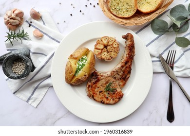 Grilled Pork Chop Steak With Baked Potato, Roasted Garlic In White Plate And Garlic Bread On The Side - Top View On Marble Background