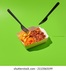 Grilled pork steak with garnish in paper box and plastic tableware. Barbecue meat with French fries and knife and fork. Fast food in minimal style on green background. Shashlik concept takeaway