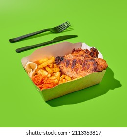 Grilled pork steak with garnish in paper box and plastic tableware. Barbecue meat with French fries and knife and fork. Fast food in minimal style on green background. Shashlik concept takeaway