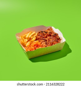 Grilled pork steak with garnish in paper box. Barbecue meat with French fries. Fast food in minimal style on green background. Shashlik concept takeaway