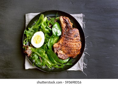 Grilled Pork Steak With Green Salad In Plate On Dark Background, Top View