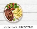 grilled pork steak marinated in spicy sauce served with mashed potato and fresh spinach tomato salad on plate on white wooden table, horizontal view from above, flat lay, free space