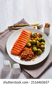 Grilled Salmon Fillet, Roast Brussel Sprouts And Boiled Farro On A White Plate On A Wooden Table, Vertical View From Above