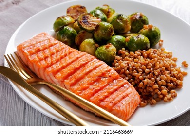 Grilled Salmon Fillet, Roast Brussel Sprouts And Boiled Farro On A White Plate  With Golden Cutlery, Close-up