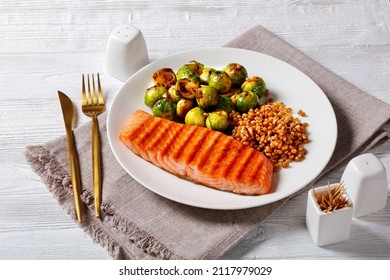 Grilled Salmon Fillet, Roast Brussel Sprouts And Boiled Farro On A White Plate On A Wooden Table, Horizontal View From Above