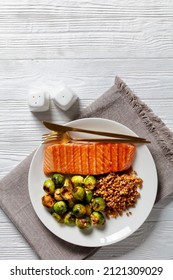 Grilled Salmon Fillet, Roast Brussel Sprouts And Boiled Farro On A White Plate On A Wooden Table, Vertical View From Above