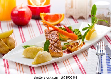 A grilled of salmon fish with mashed potatoes, caramelized onion, carrot, broccoli, bell pepper and lemons on white plate. An apple, orange, red ramekin bowl, a white lantern and a jar of pepper.