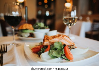 Grilled salmon with mixed salad and avocado cheese burger sitting on table with white table cloth in fine dining restaurant with window light. Film grain look