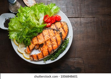 Grilled salmon steak served with grilled tomatoes, salad, lemon sliced, pepper seeds, rosemary, parsley on white plate on wooden table.