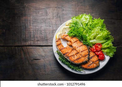 Grilled salmon steak served with grilled tomatoes, salad, lemon sliced, pepper seeds, rosemary, parsley on white plate on wooden table.