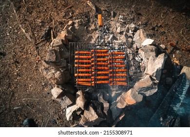 Grilled Sausages Are Fried In A Grid. There Is A Bonfire In The Tourist Camp. Beautiful Decoration Paved With Stone. The Concept Of Delicious Food On A Hike. Top Down View.