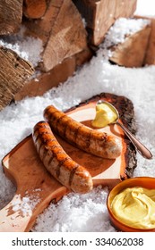 Grilled sausages served on a rustic wooden board with a scoop of mustard outdoors in the snow at a winter grill party