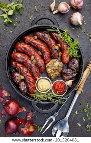 Grilled sausages and vegetables with the addition of ketchup and mustard served in a black cast iron dish, top view