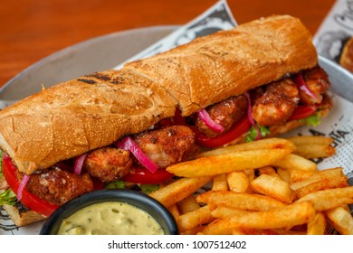 Grilled shrimp sandwich with sauce and spices, tomato, lettuce on a wooden table with French fries and white onion sauce.