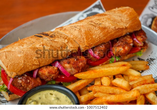 Grilled\
shrimp sandwich with sauce and spices, tomato, lettuce on a wooden\
table with French fries and white onion\
sauce.