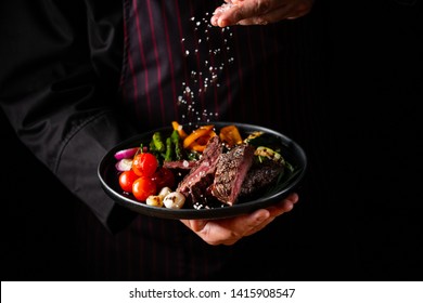 Grilled And Sliced Beef Steak With Grilled Vegetables Served On Black Plate On Black Background Presentation In Chef Hands.