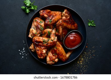 Grilled spicy chicken wings with ketchup on a black plate on a dark slate, stone or concrete background. Top view with copy space.