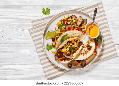 Grilled Steak Tacos with olives, tomatoes, red onion, avocado, cilantro, corn, shredded cheddar cheese and thousand island sauce on platter on white wood tabe, flat lay, free space - Powered by Shutterstock