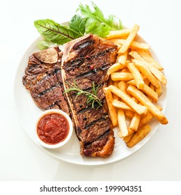 Grilled t-bone or porterhouse steak seasoned with rosemary and served with golden French fries, fresh leafy herb salad and a tomato dip, high angle view on white