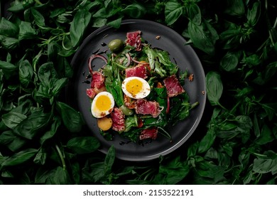 Grilled Tuna Slices With Fried Lettuce Leaves, Vegetables, Olives And Sesame Seeds In A Plate On A Background Of Basil Leaves. Asian Fish Dish Of Tuna Fillet