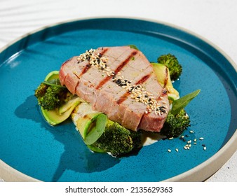Grilled tuna steak with broccoli and zucchini in modern ceramic plate. Healthy food - roasted tuna with green vegetables. Fish dish in minimal style. Tuna fillet in blue plate with hard shadow