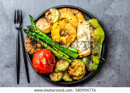 Grilled vegetables - tomatoes, asparagus, zucchini, bell pepper, pineapple slices and garlic on cast iron black plate, gray slate background. top view