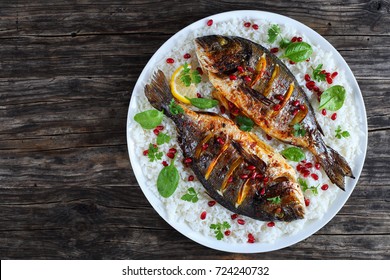 Grilled Whole Fish Loaded With Fresh Citrus, Herbs Spices And Pomegranate Seeds On Bed Of Rice, On White Plate On Old Dark Wooden Table, View From Above