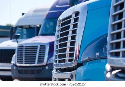 Grilles Of Different Models Of Big Rig Semi Trucks Which Stand In Row On The Truck Stop Parking Waiting To Continue Delivery Cargo 