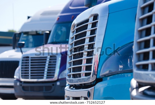 Grilles of different models of big rig semi trucks which\
stand in row on the truck stop parking waiting to continue delivery\
cargo 