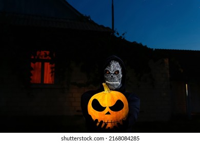 Grim Reaper Holding Halloween Pumpkin Head. Man In Death Mask With Fire Flame In Eyes On Night Sky, Light Window Background. Halloween Holiday Concept. Dark Horror.