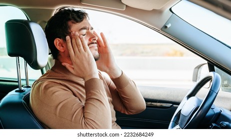 A grimace of pain on the face of a young male driver sitting at the wheel of a car, hands on his temples. He closed his eyes, held his head in his hands, let go of the steering wheel.