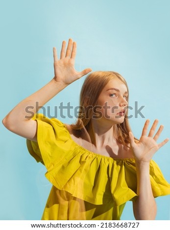Grimace. Young pretty girl, student leaned against transparent glass isolated on light blue color background. Close-up. Concept of human emotions, facial expression, music, ad, fashion, beauty.