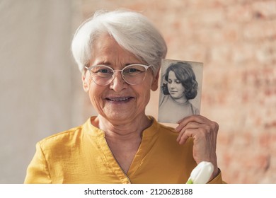 Grinning And Content Grey-haired Grandma Comparing Herself To Her Old Black And White Portrait . High Quality Photo